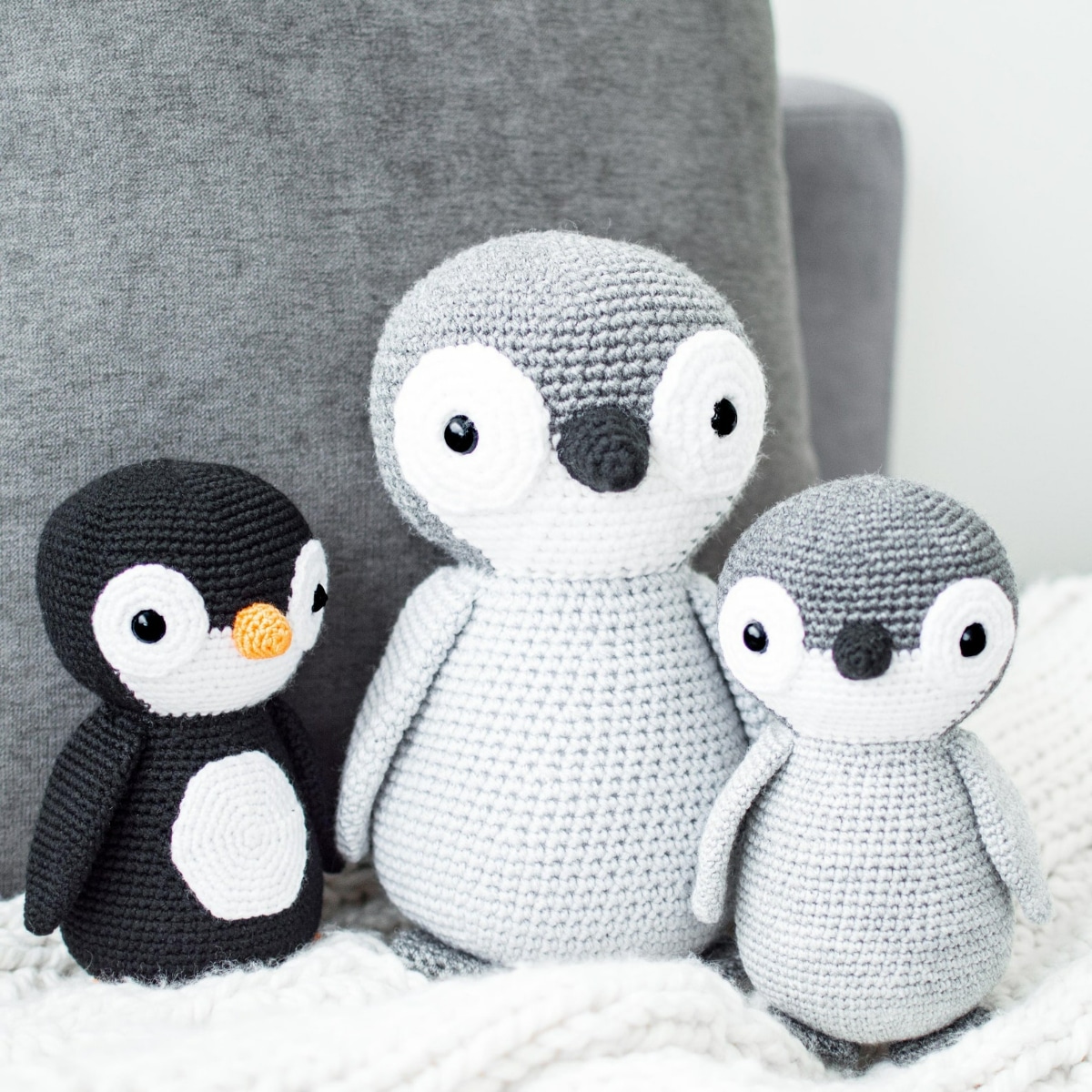 Large grey crochet penguin with a smaller gray penguin and a black and white penguin standing on a white blanket.