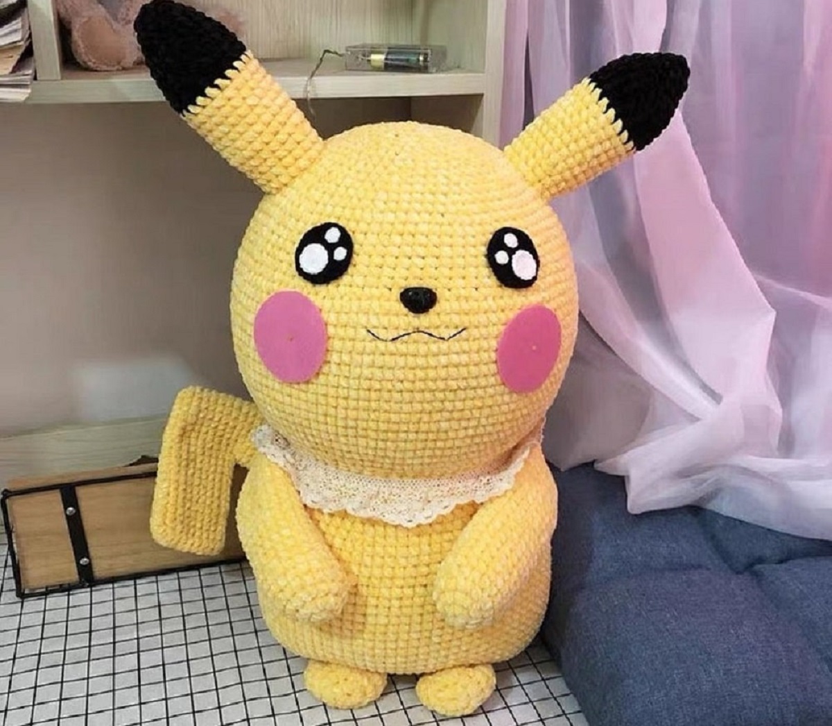 Large stuffed crochet Pickachu with small black and white eyes standing on a table and wearing a white lace collar. 