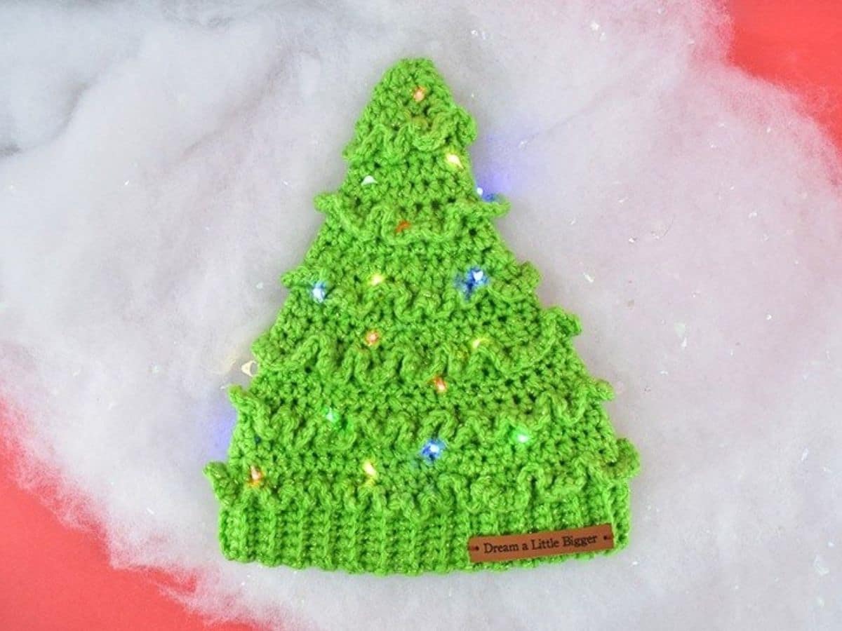A green crochet Christmas tree style hat with small yellow and pink bobbles stitched in lying on fake snow. 