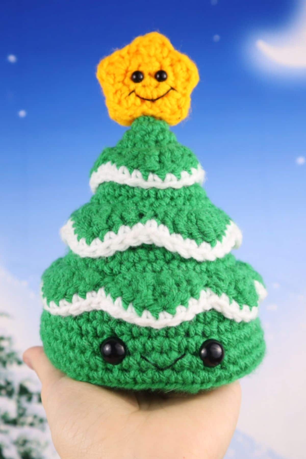 A hand holding a green crochet Chrismtas tree with eyes at the base and a yellow star on top smiling.