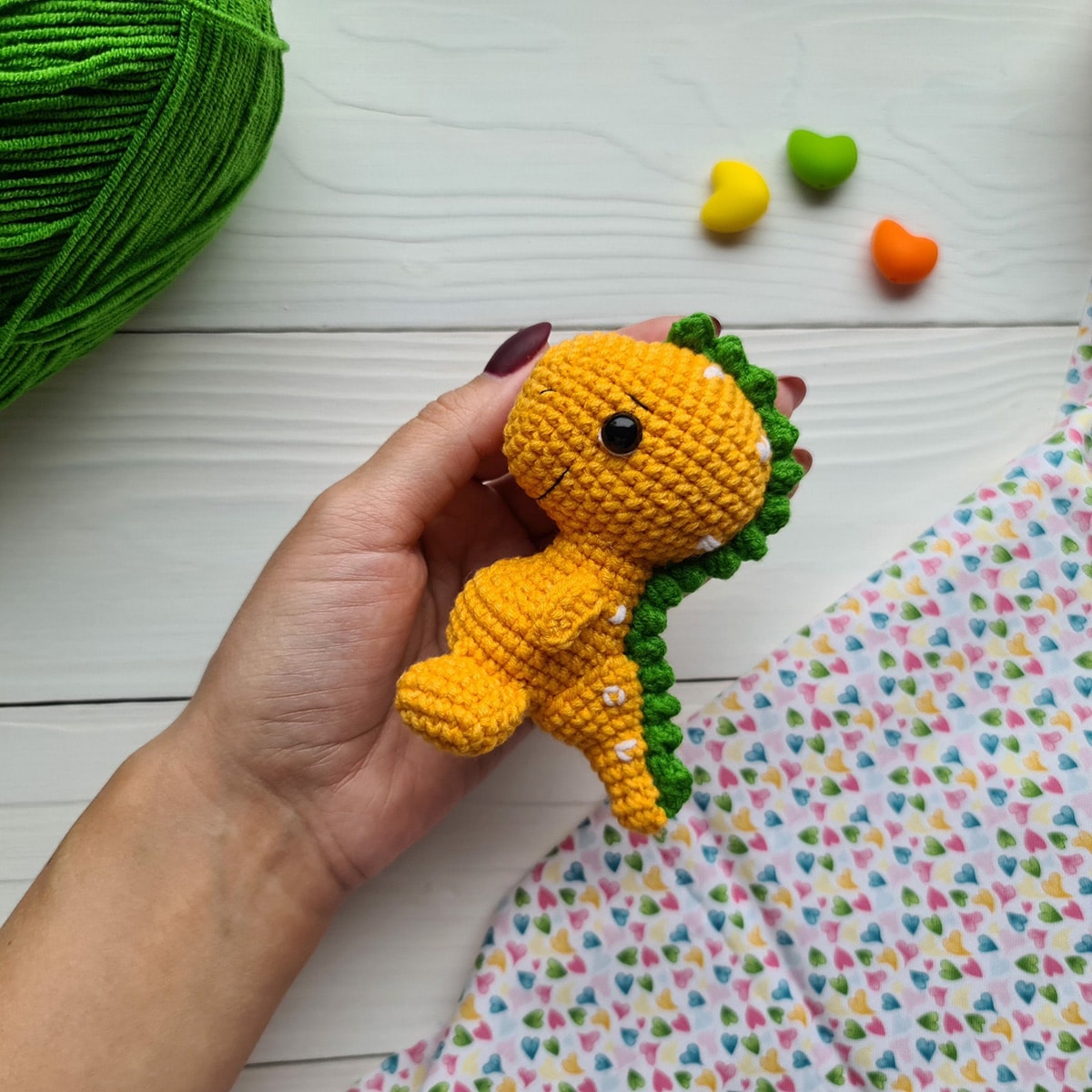 A manicured hand holding a small yellow crochet baby dinosaur with green spikes down its back on a white wooden background.