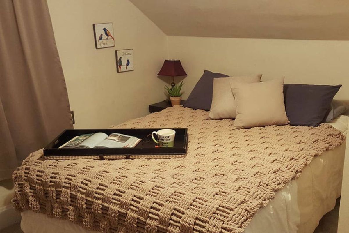 A bed with several throw cushions and a beige crochet blanket with a chunky basketweave pattern spread over it.