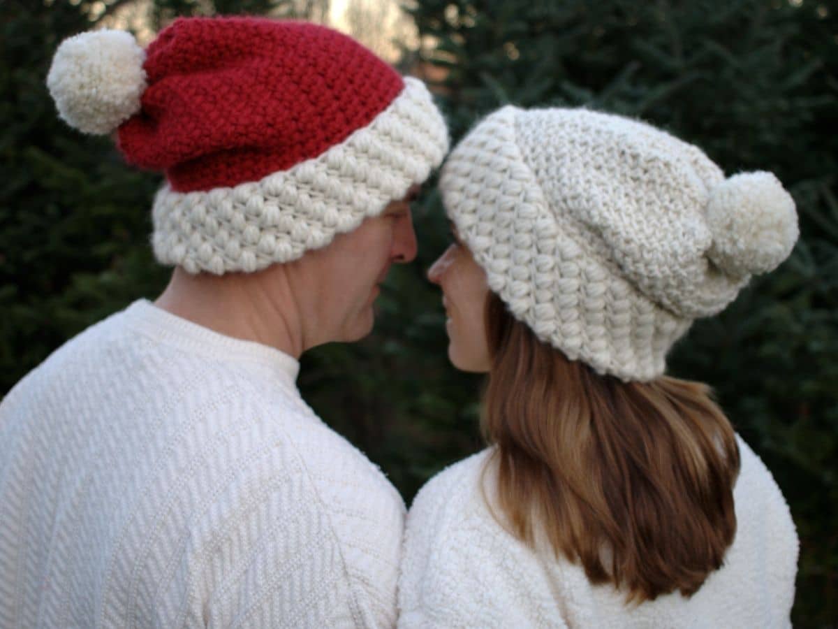 Man and woman in white jumpers standing next to each other in Santa hats. The man’s hat is red with a white bobble and the woman’s white.