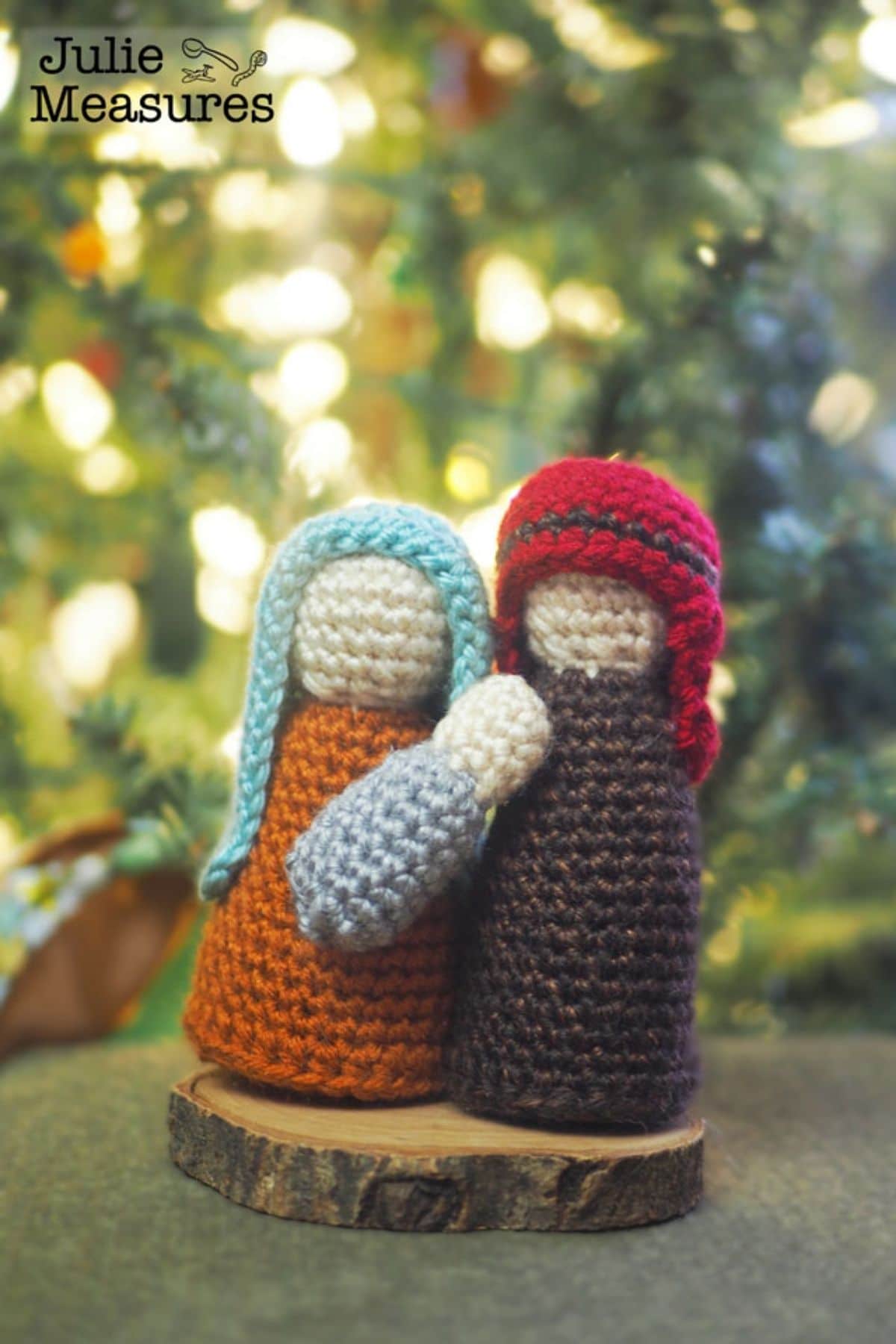 Faceless crochet Mary and Joseph standing on a small wooden log holding a small baby Jesus in their arms.