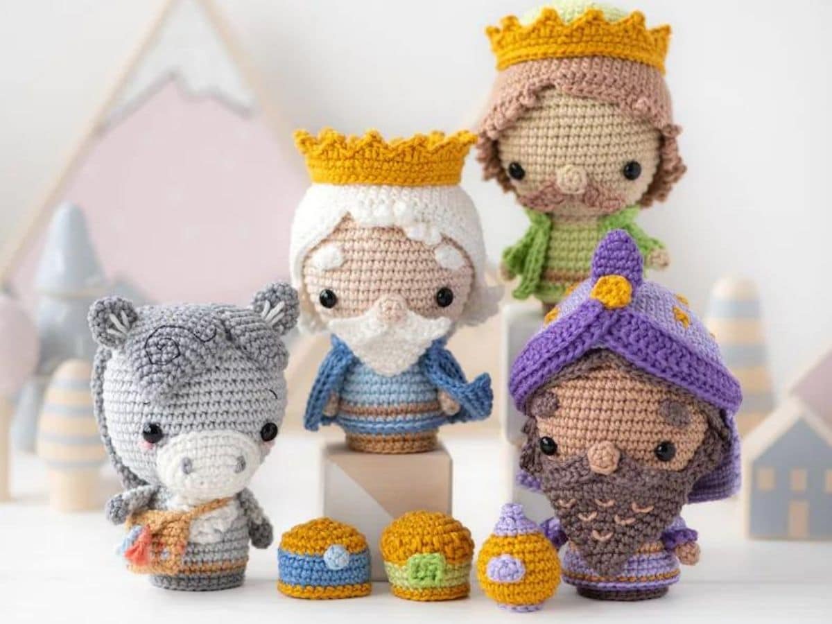 Three wise men crochet dolls standing next to a donkey with crochet trinkets in front of them and a pale pink and white background.