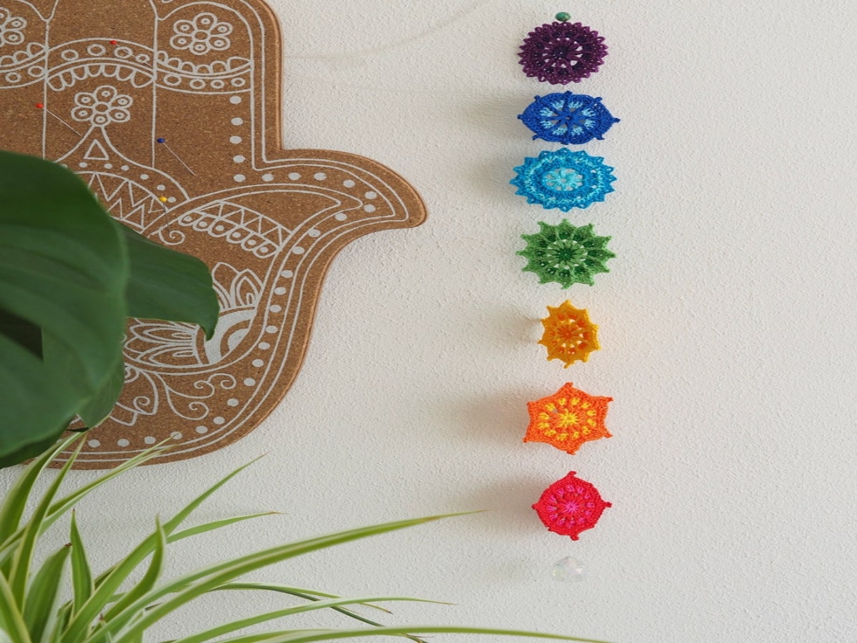 A small crochet wall hanging of mini mandalas in purple, plue, green, orange and pink on a cream wall next to a plant.