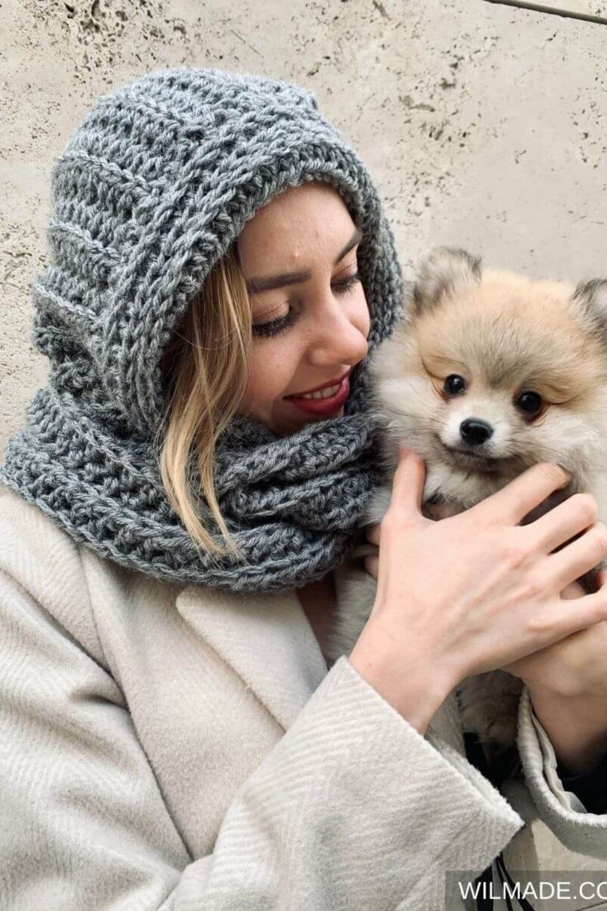 Blonde woman holding a Pomeranian wearing a light gray crochet hooded scarf over her head and neck.