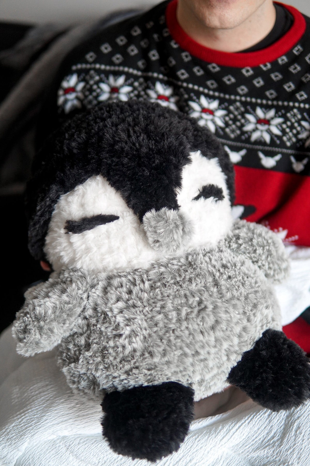 Large crochet penguin with a black and white face and gray fluffy body held by someone wearing a black and red Christmas jumper.