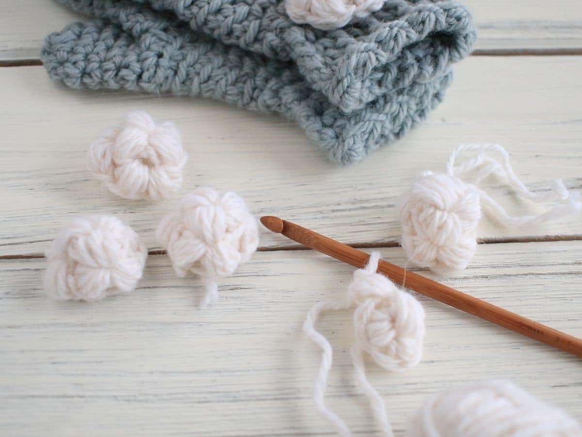 Small white snowballs on a white wooden table. One ball is still on the knitting needle sat next to pale blue gloves. 