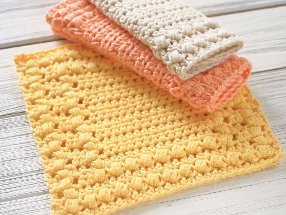 Yellow crochet dishcloth square with orange and cream cloths folded on top in the right corner sitting on a pale wooden table. 