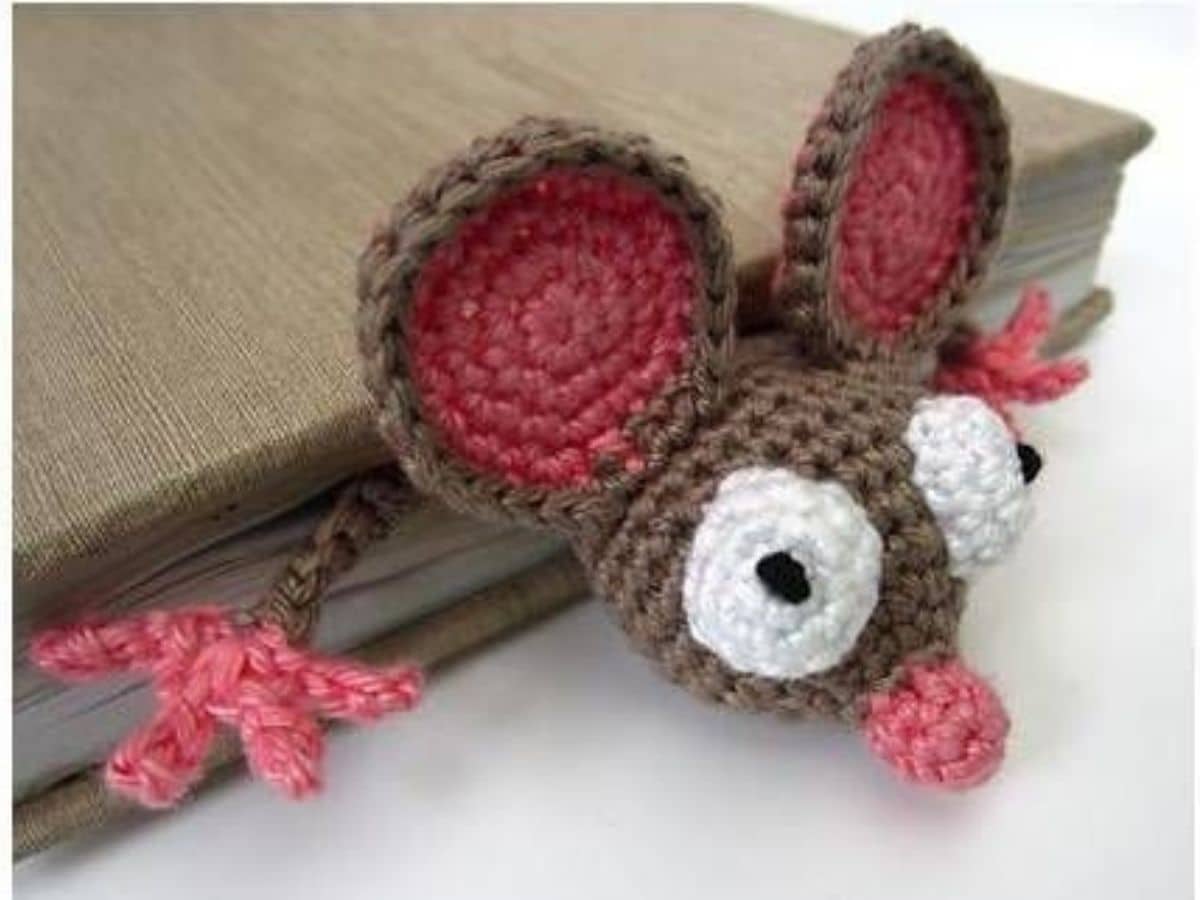Crochet brown mouse bookmark with its face, ears, and hands sticking out of a brown book on a white background. 