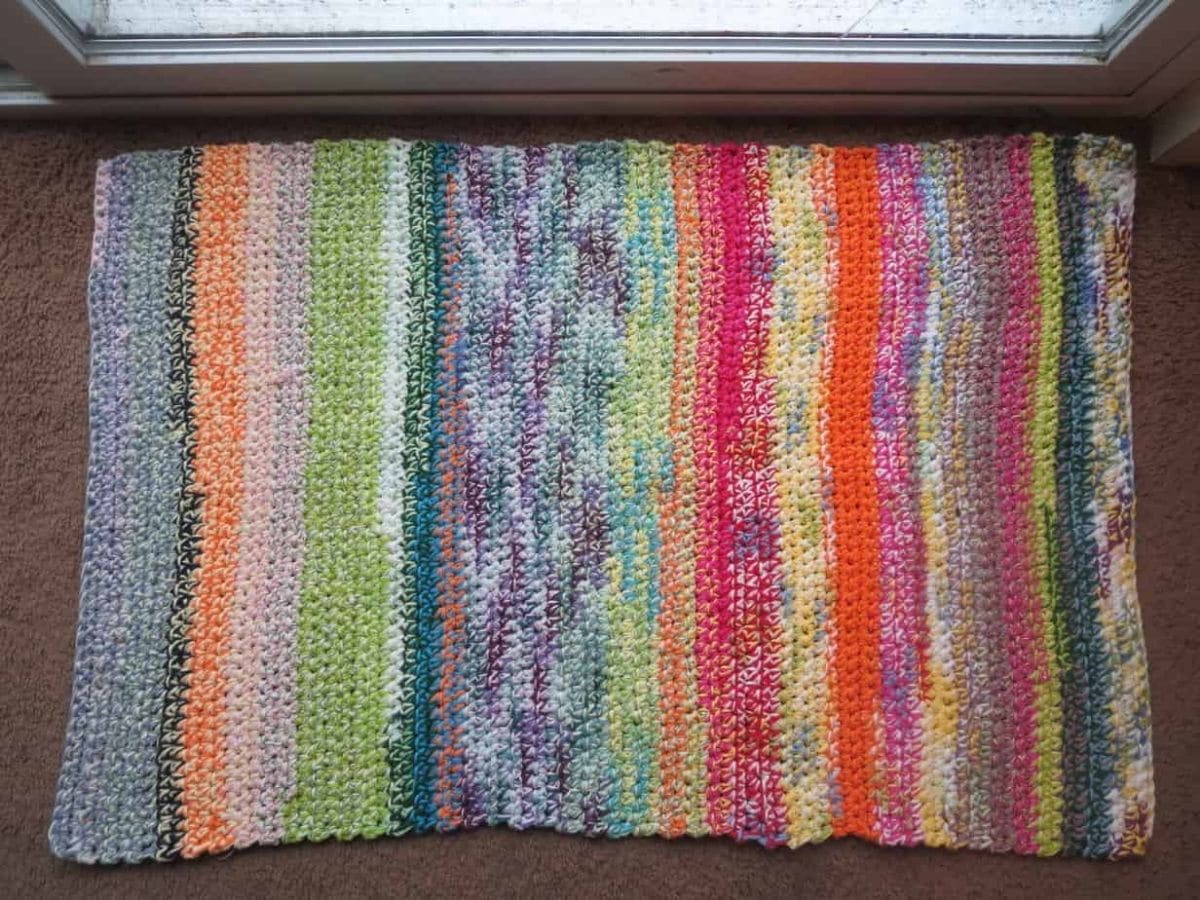 Multi colored crochet rug with pink, red, purple, green, and orange vertical stripes lays on a brown floor by a glass door. 