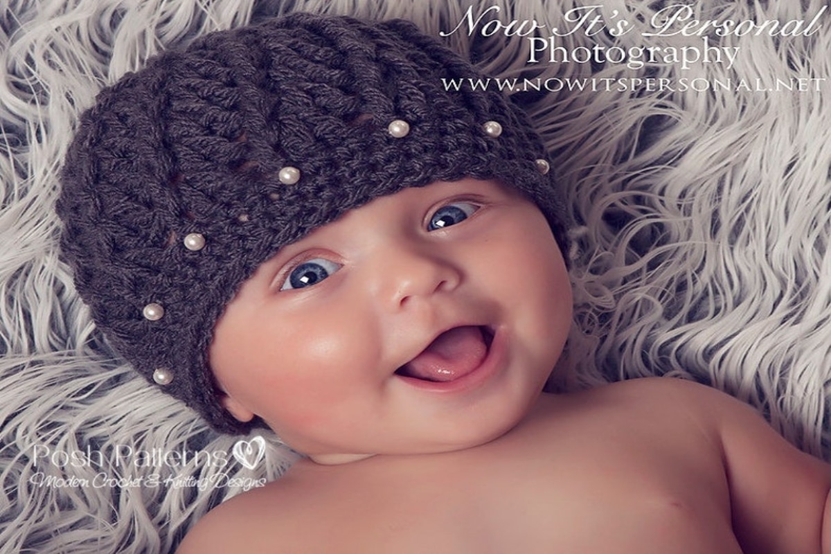 A smiling baby wearing a purple crochet beanie hat with small pearls stitched into the banding.