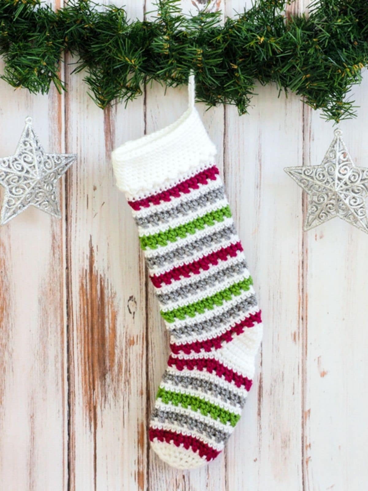 White, red, green, and gray striped crochet Christmas stocking hanging from pine with two star decorations on a pale wooden background. 