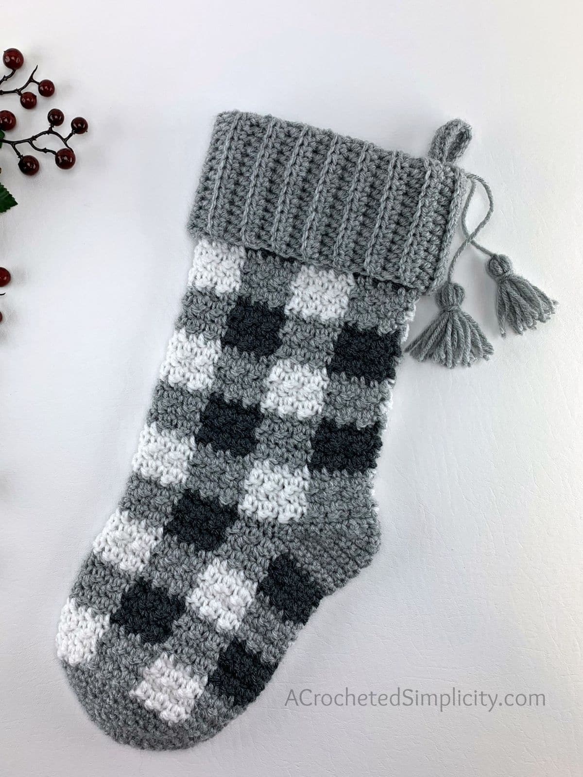 Black, white, and gray plaid crochet stocking with two gray tassels on the top laying on a white table with some holly offside. 