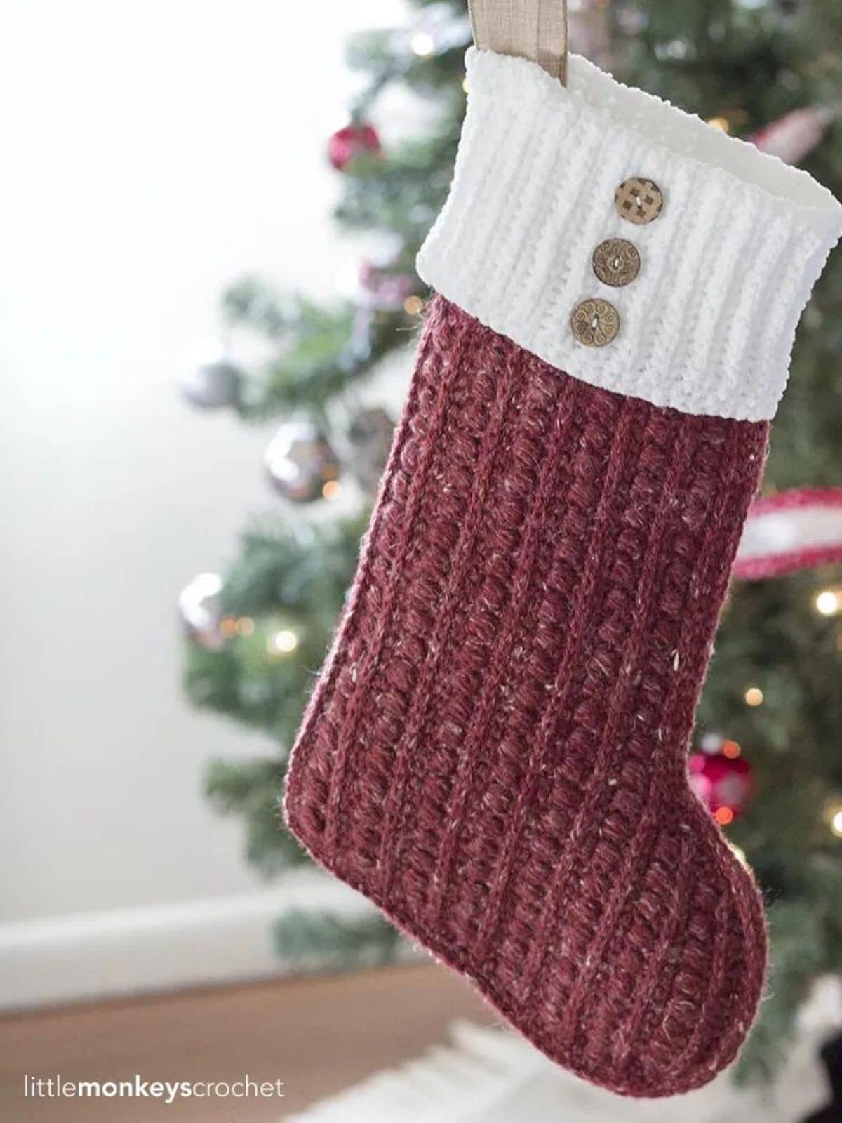 Red chunky knit stocking with white banding and three brown decorative buttons hanging in front of a decorated Christmas tree. 
